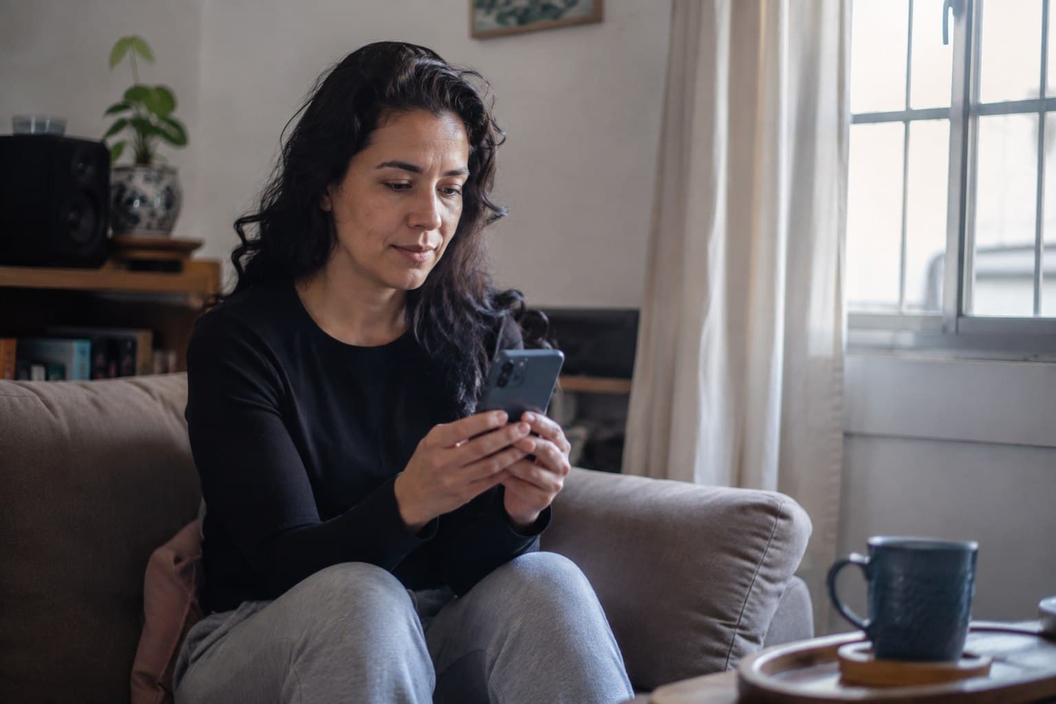 Mulher sentada no sofá de casa, olhando o celular com calma.
